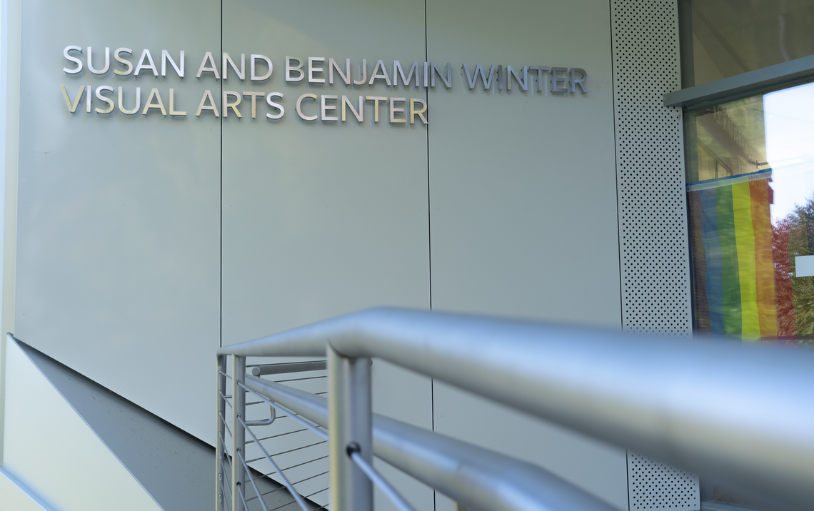 The ramp entrance to Winter Visual Arts Center. "Susan and Benjamin Visual Arts Center" is written on the wall in silver. In the window next to the glass doors, a gay pride flag is visible.