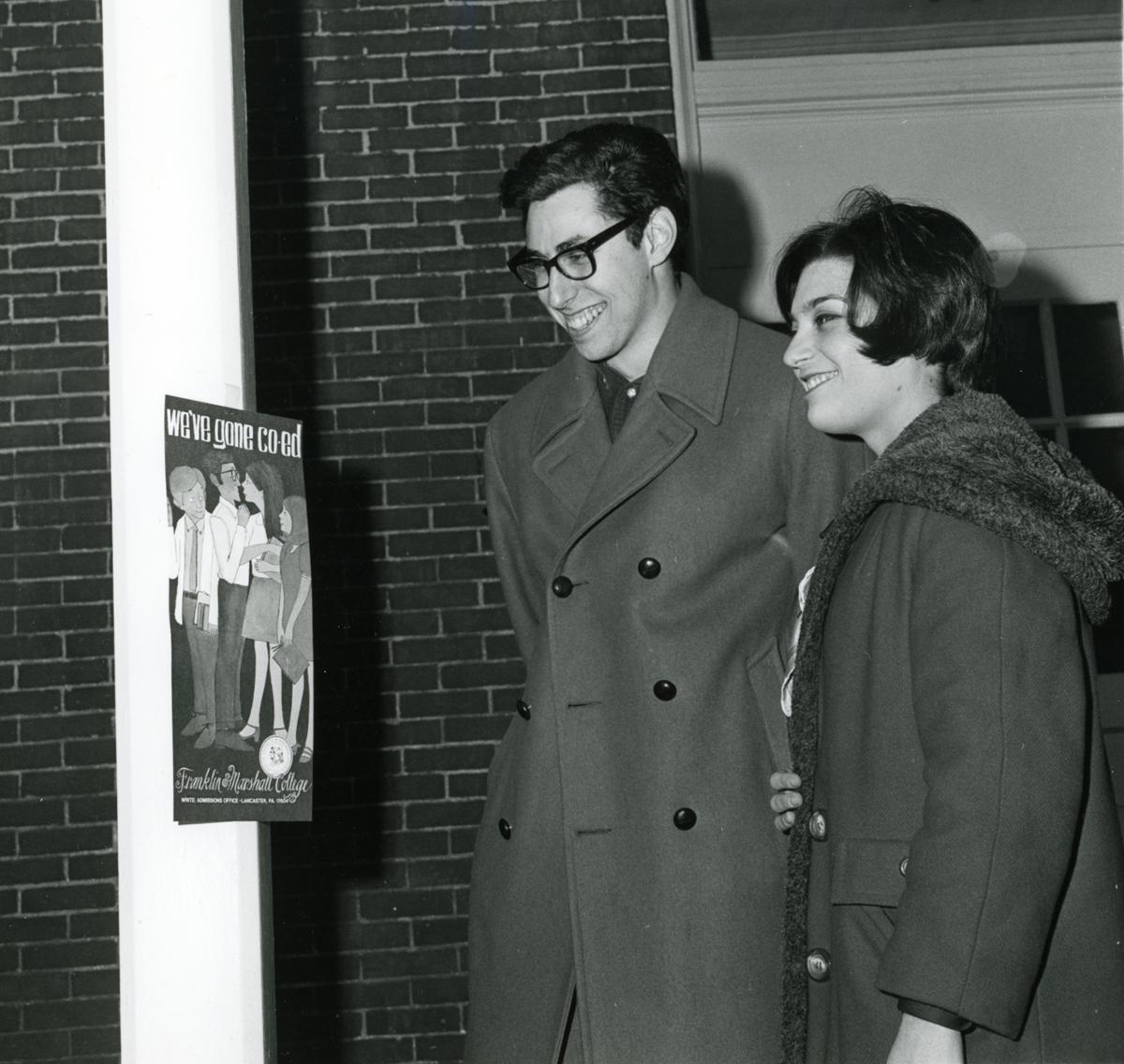 Two students looking at a poster reading "We've gone coed." Notes on the verso suggest that the photograph was printed in an F&M publication.